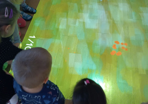 Dzieci siedzące dookoła magicznego dywanu (interaktywna zabwka, która wyświetla na podłodzę gry dla dzieci)