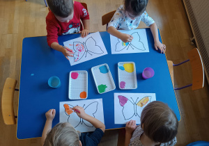Czworo dzieci widoczne z góry siedzą przy niebieskim stoliku i malują farbami szablon motyla.