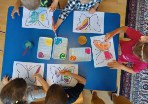 Pięcioro dzieci widoczne z góry siedzą przy niebieskim stoliku i malują farbami szablon motyla.