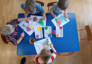 Czworo dzieci widoczne z góry siedzą przy niebieskim stoliku i malują farbami szablon motyla.