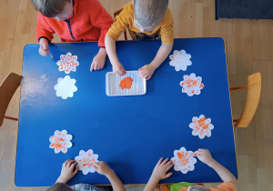Dzieci siedzące przy stoliku. Widziane z góry. Stemplują kwiatki wycięte z papieru przy użyciu patyczków higienicznych.